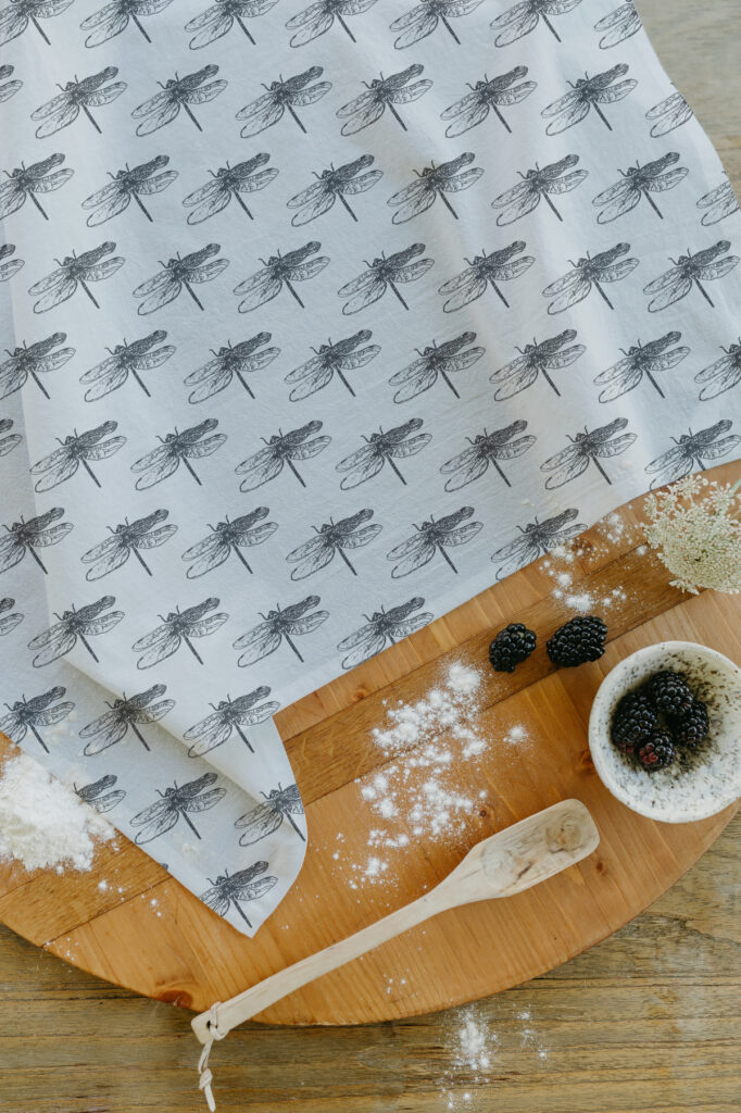 A dragonfly patterned tea towel resting on a wooden cutting board alongside a wooden spoon and some blackberries.